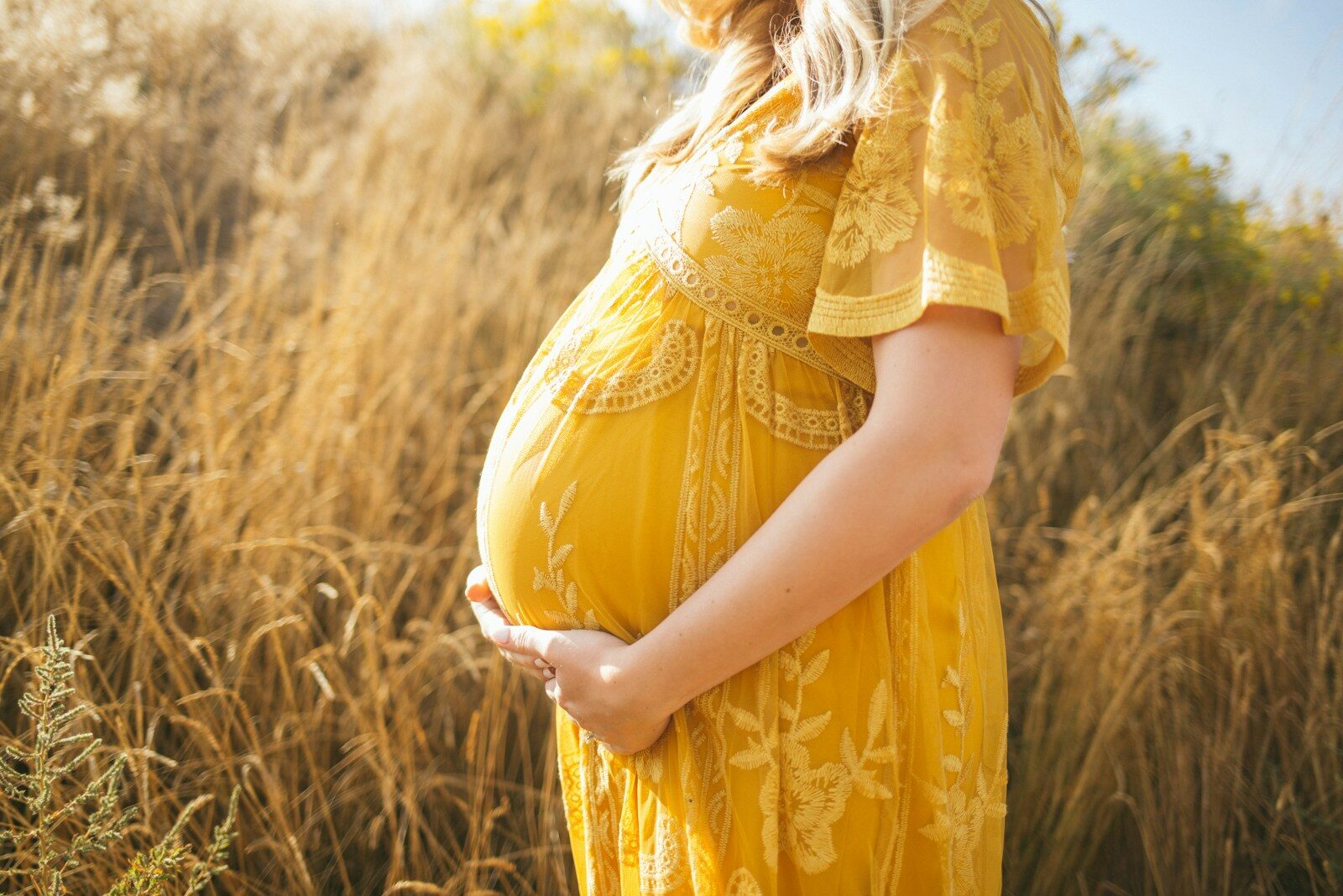 pregnant woman wearing yellow floral dress standing while touching her tummy and facing her right side near brown field during daytime relacionada a Contribuir depois da gravidez garante salário-maternidade?